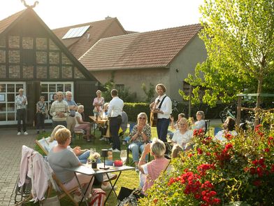 Menschen sitzen auf einer Wiese bei einem Picknickkonzert in Hille, umgeben von Bäumen und Fachwerkhäusern.