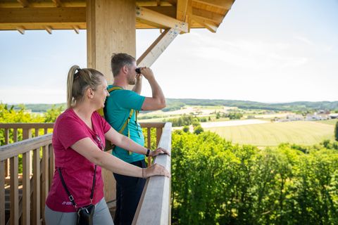Zwei Personen stehen auf einem Aussichtsturm und genießen den weiten Blick über eine grüne Landschaft.