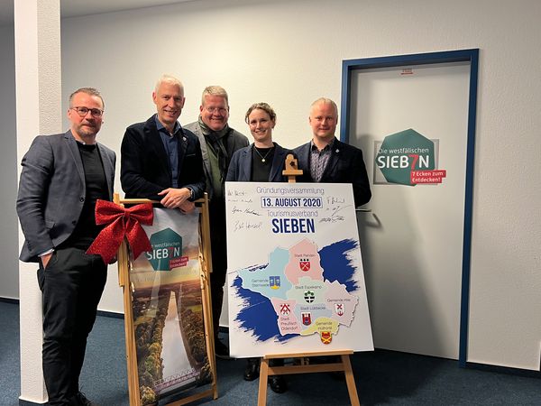 Gruppe von Personen in Innenraum neben zwei Schautafeln auf Staffeleien und einer Tür mit dem Logo der „Westfälischen Sieben“.