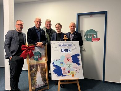 Gruppe von Personen in Innenraum neben zwei Schautafeln auf Staffeleien und einer Tür mit dem Logo der „Westfälischen Sieben“.