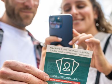 Personen betrachten eine Broschüre zur Siebenecken-App, während sie mit dem Smartphone interagieren.