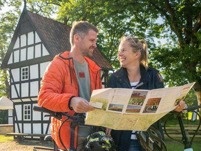 Zwei Personen mit Fahrrädern stehen vor einem historischen Fachwerkgebäude und betrachten eine Karte.