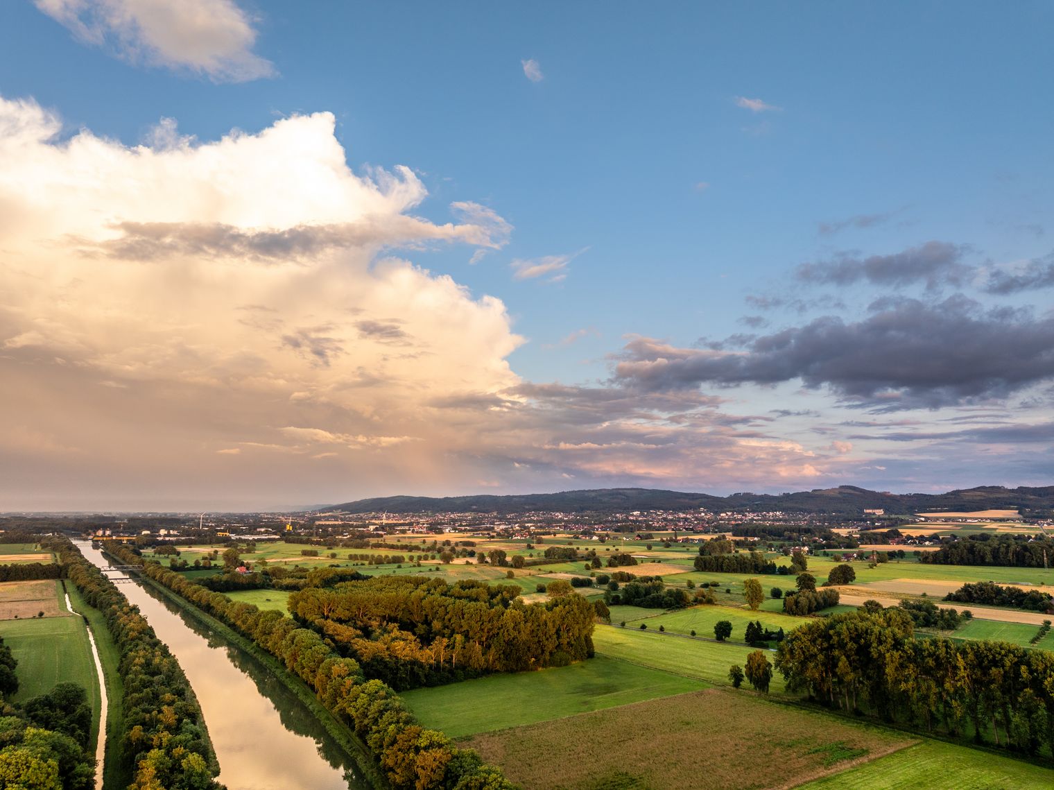 Weitläufige Landschaft mit Feldern, Bäumen und dem Mittellandkanal, der sich gerade durch die Szenerie zieht; darüber ein Himmel mit hellen Wolken und dunkleren Wolkenfeldern in der Ferne.