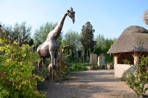 Metallskulptur einer Giraffe mit umliegender Vegetation und einem Strohdachpavillon im Hintergrund.